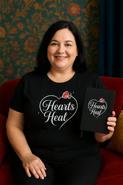 Woman wearing a black t-shirt with 'Hearts Heal' design, sitting on a couch, holding a matching notebook.