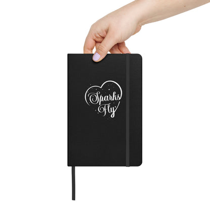 Black hardcover Sparks Fly Heart notebook held by a person.